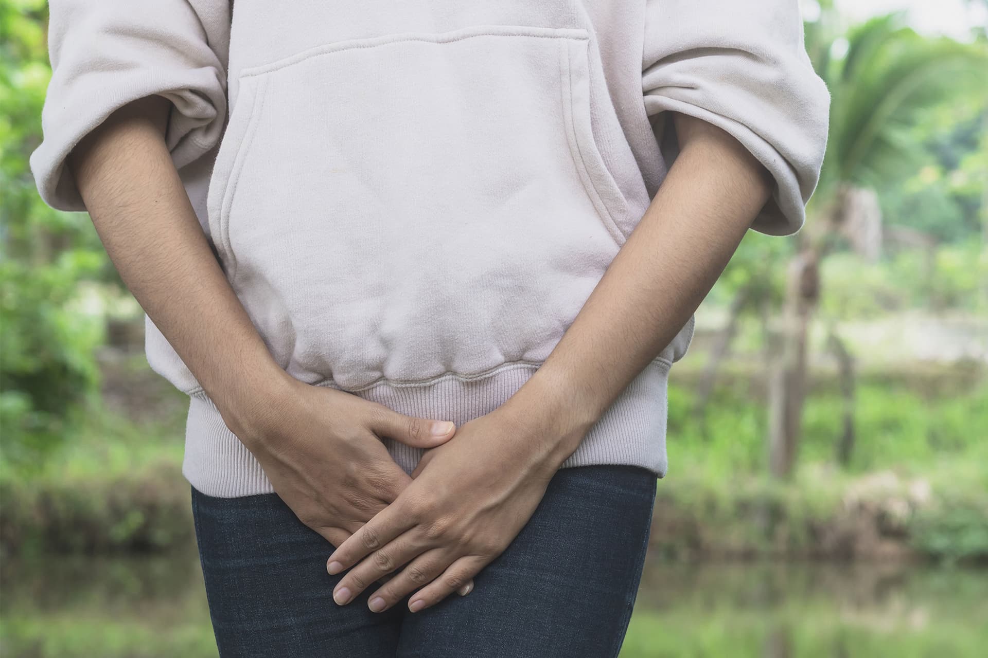 Person outdoors with hands clasped over their lower abdomen, possibly indicating pelvic pain.