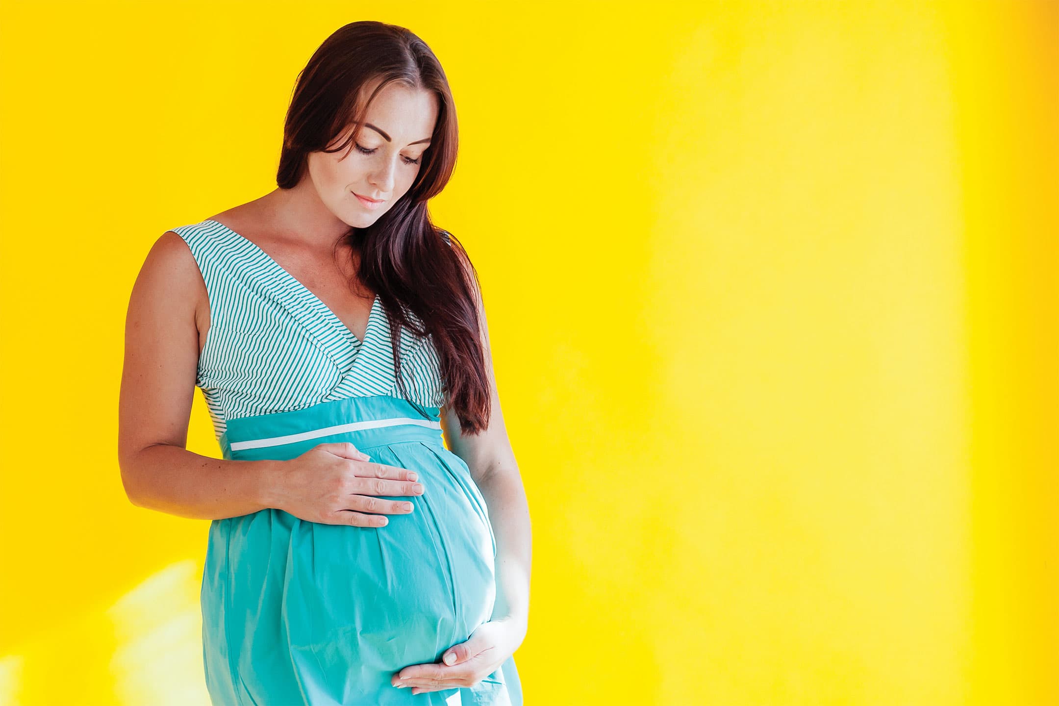 A pregnant woman with long dark hair stands against a bright yellow background, looking down and cradling her baby bump.