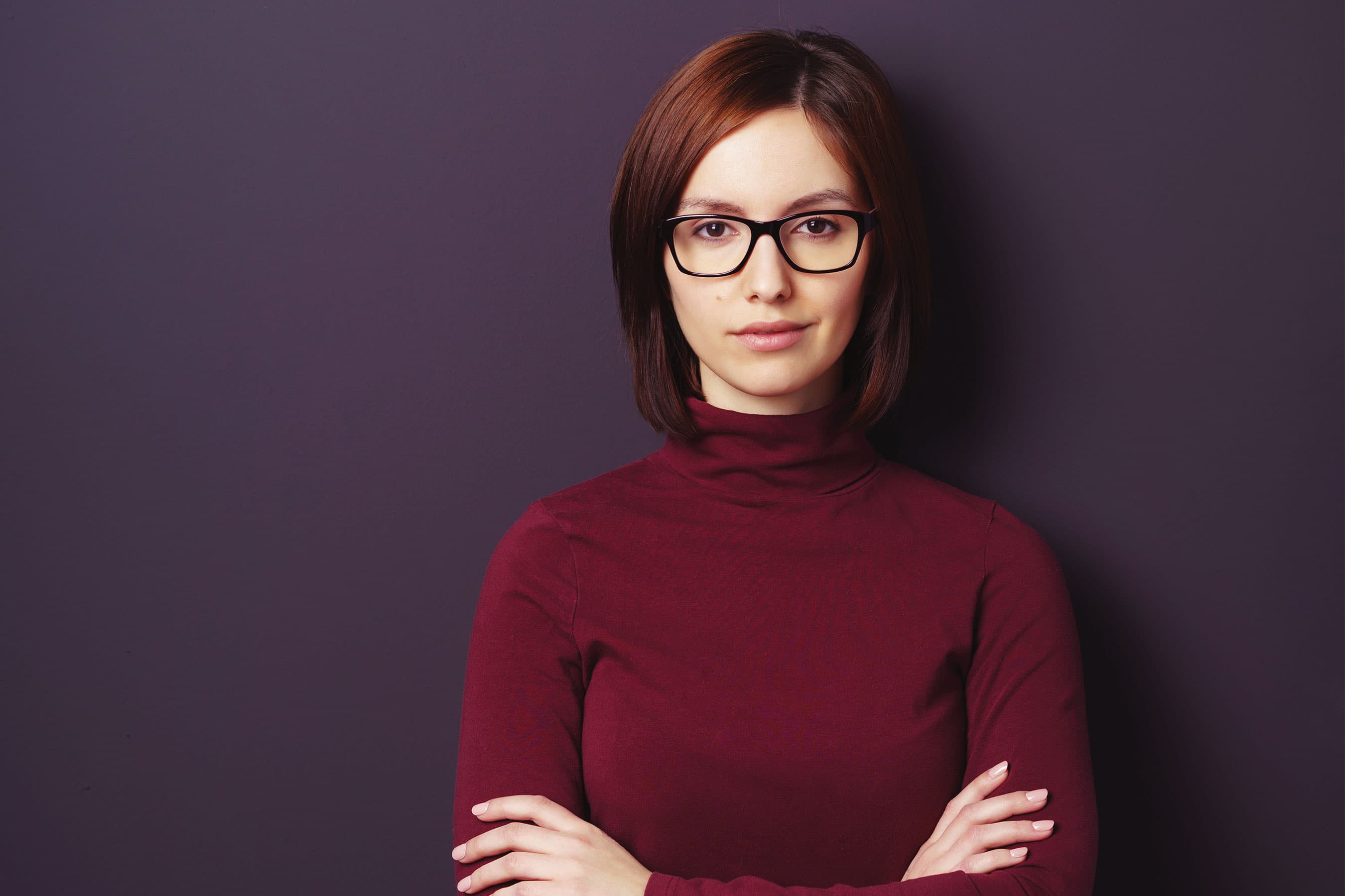 Serious woman in glasses standing with arms crossed against a dark purple background.