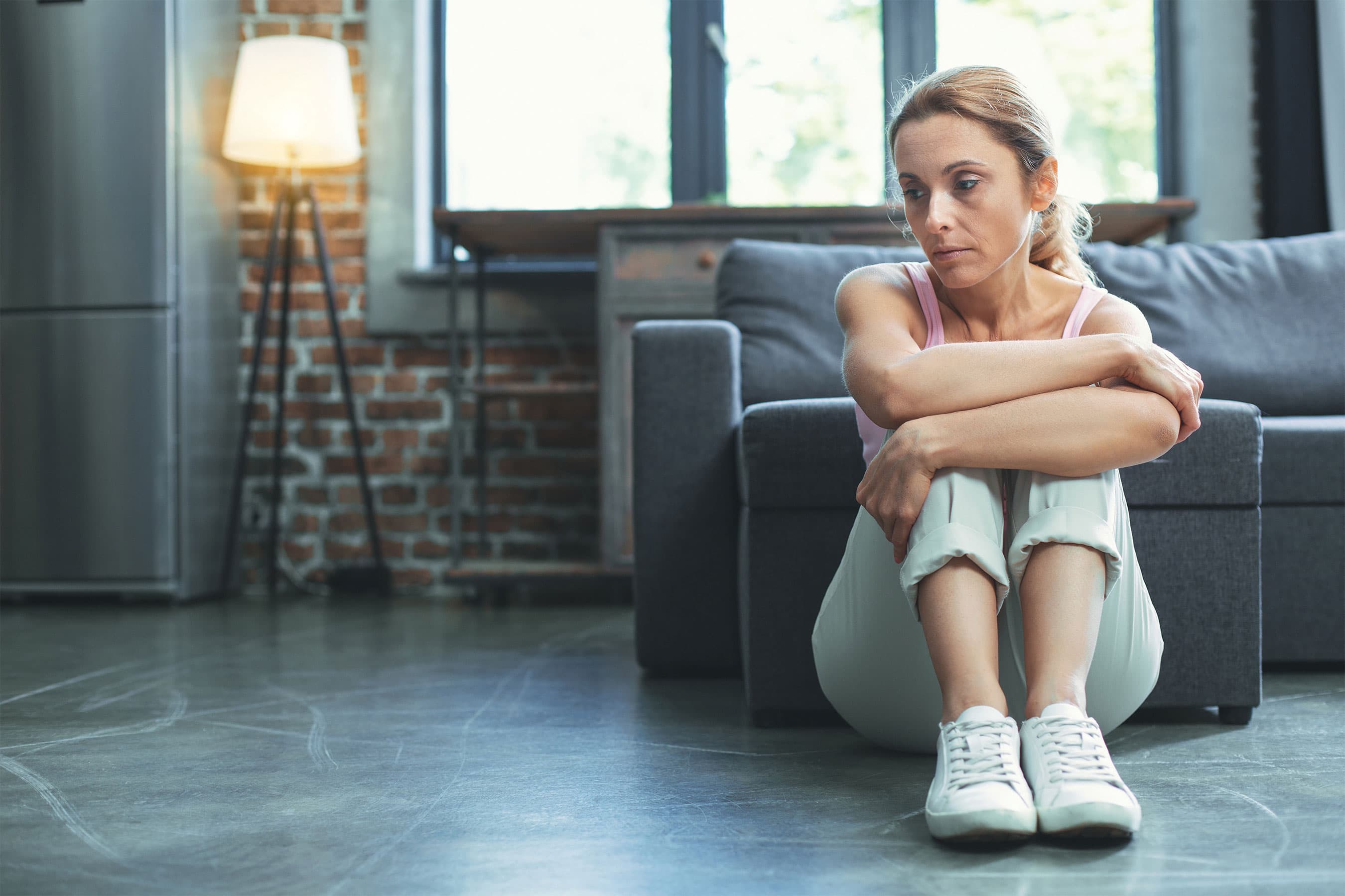 Woman sitting on the floor near a couch, hugging her knees and looking uncomforable.