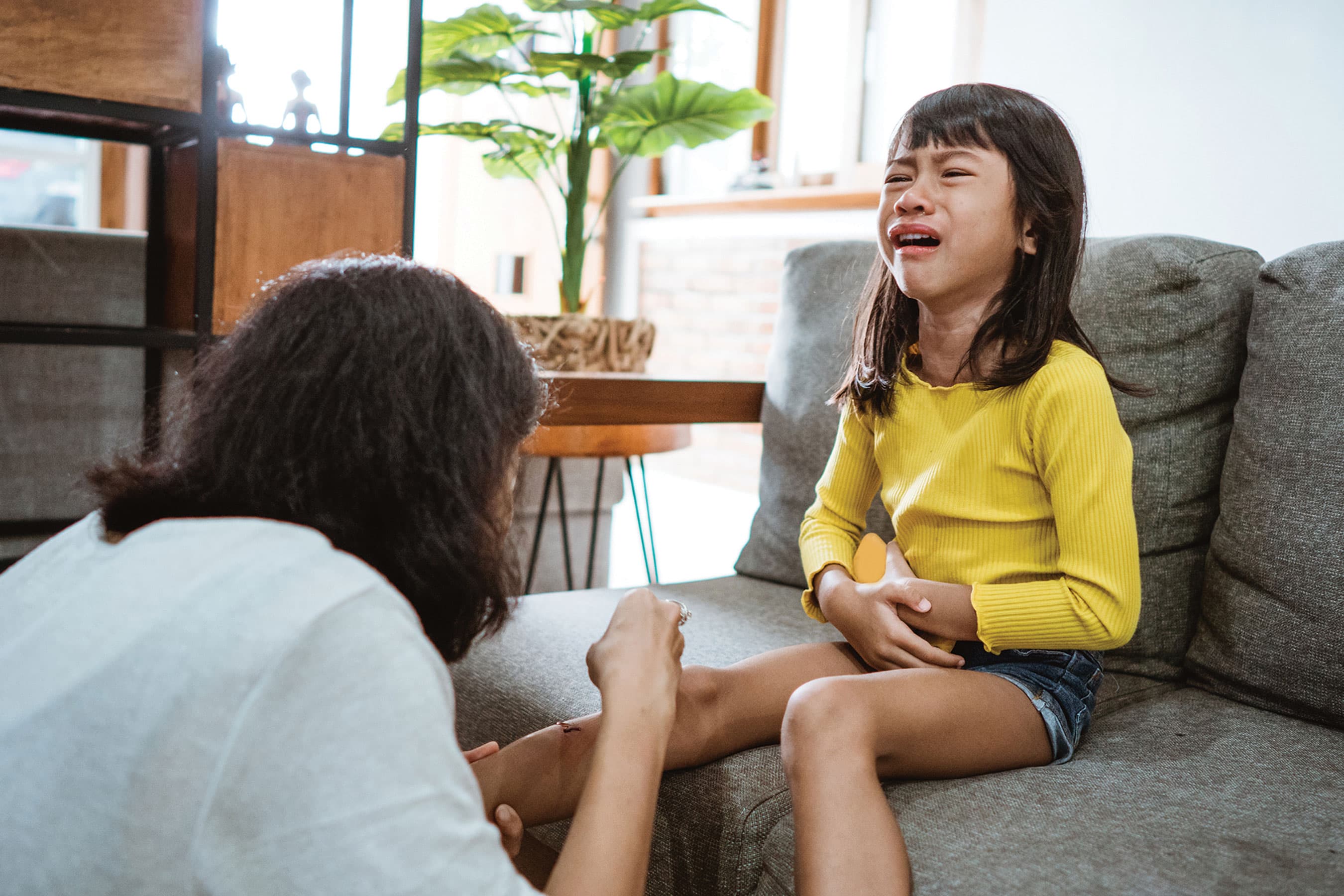 A mother gently tends to a scrape on her crying daughter's knee while she sits on a sofa.